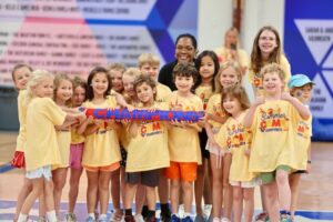 Summer Camp-1 2025 Spirit Stick winners Sloane (red skirt), Caroline (white shorts), Morgan, Neely (pink shorts), Maeve (orange shorts), Sha, Piper, Jaiden (boy in grey shorts), Macklin (boy behind), Simon (boy in orange shorts), Coach Chelsea Tubonemi, Spring (girl in black shorts), Sloane (girl in pink shorts), Isabella (girl in tie dye shorts), William (boy in green shorts), Ingrid (girl in blue hat), and Ryan Elizabeth (behind, long hair)